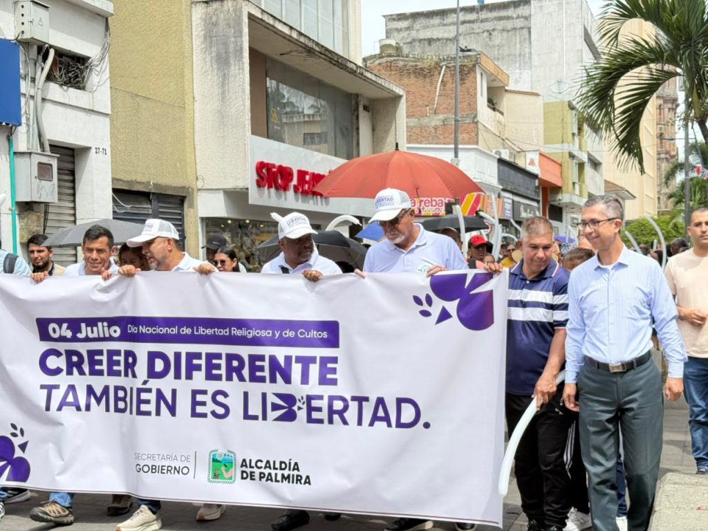 Fotografía de una marcha en Palmira con motivo del Día Nacional de la Libertad Religiosa y de Cultos. Un grupo de personas camina sosteniendo una gran pancarta blanca con logotipos de la Alcaldía de Palmira y la Secretaría de Gobierno, en la que se lee: "Creer diferente también es libertad".