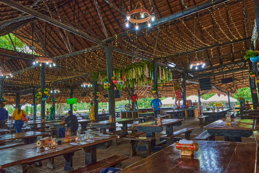 Fotografía del amplio comedor interior de un restaurante campestre en Palmira, con arquitectura tradicional de techo de paja y estructuras de madera. Se aprecian mesas largas, luces decorativas en el techo y algunas personas comensales y trabajadoras en el fondo.