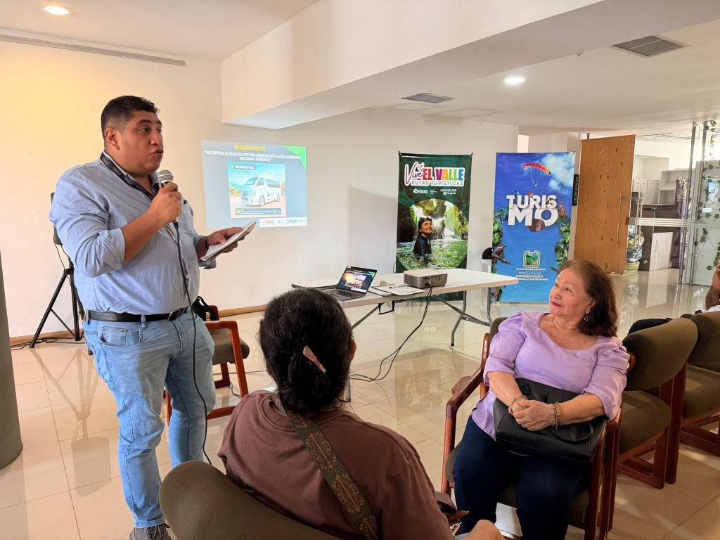 En la foto un facilitador expone con micrófono en mano frente a un grupo de personas que asisten a un taller de turismo local en un salón iluminado.