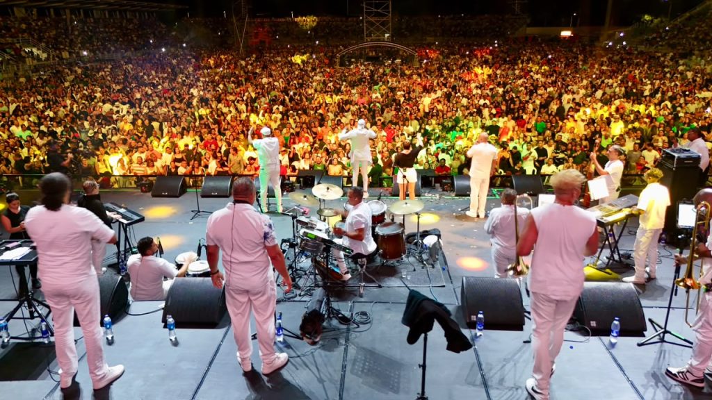 En la fotografía una agrupación musical vestida de blanco actúa en un gran escenario frente a una multitud en un concierto nocturno al aire libre en Estadio Rivera Escobar.