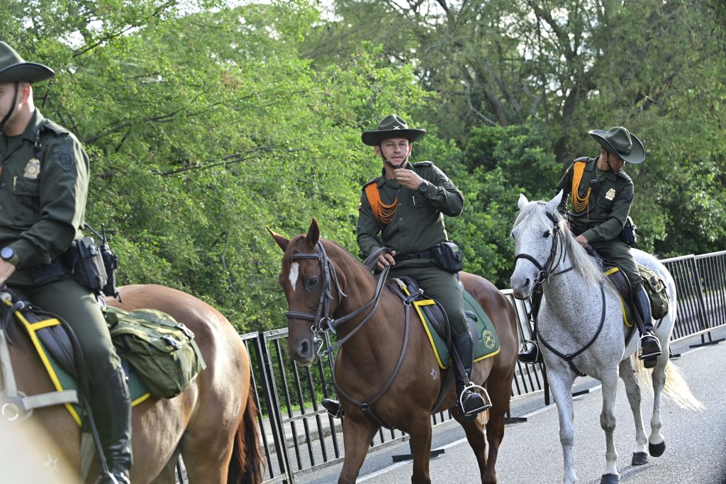 En la fotografía tres integrantes de la policía montada patrullan a caballo por una vía asfaltada junto a una zona verde. Las personas visten uniformes de color verde oliva, sombreros de ala ancha y cordones ornamentales en los hombros. Dos montan caballos castaños y una monta un caballo tordo blanco.