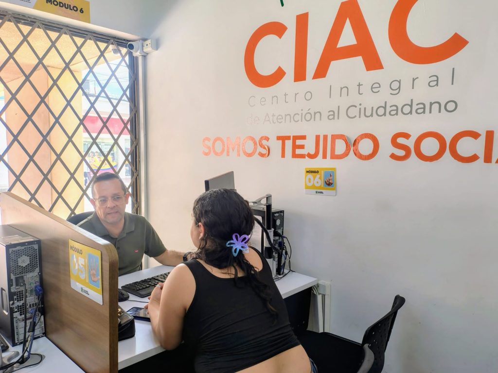 En la fotografía una persona en su puesto de trabajo atiende a una ciudadana en una oficina pública de servicio. En la pared del fondo se lee un letrero institucional que dice Centro Integral de Atención al Ciudadano.