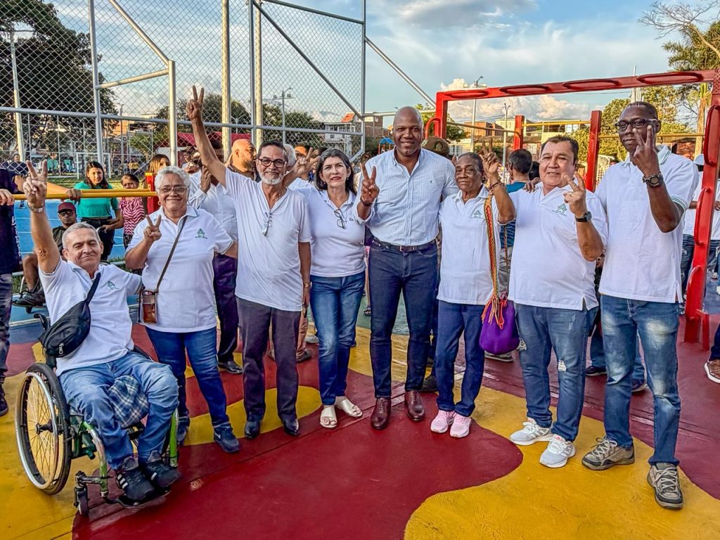 Una imagen un grupo diverso de personas sonríe y hace la señal de la victoria en una cancha deportiva al aire libre. Entre ellas hay una persona usuaria de silla de ruedas. La mayoría viste camisetas blancas y pantalones de mezclilla.