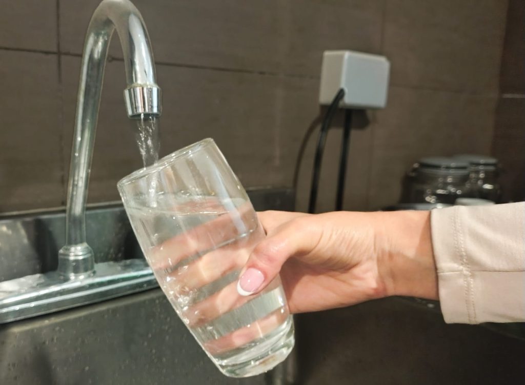 Fotografía en primer plano de una mano sosteniendo un vaso de vidrio que se llena con agua directamente de un grifo de cocina.