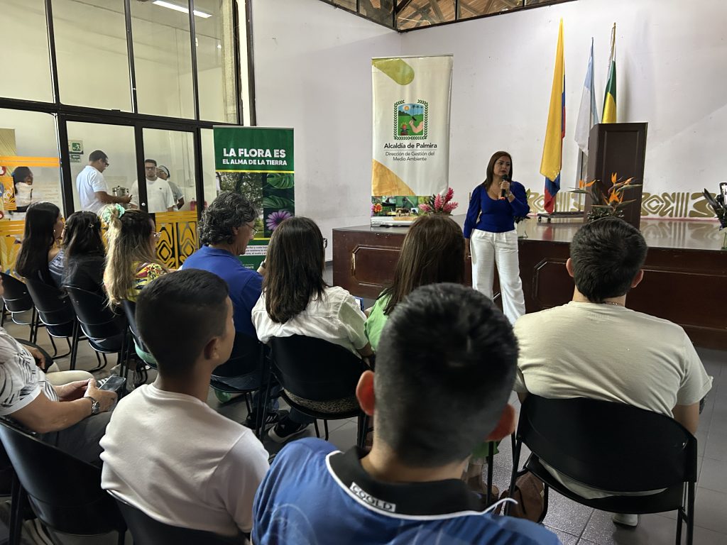 Una funcionaria realiza una exposición ante un grupo de personas en un auditorio. Al fondo se observan banderas y un pendón de la Dirección de Gestión del Medio Ambiente de la Alcaldía de Palmira.