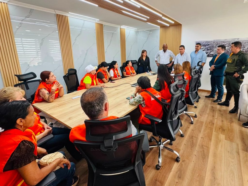 En la fotografía se ve una reunión en una sala de juntas donde un grupo de personas con chalecos naranjas está sentado alrededor de una mesa de madera, escuchando atentamente a un equipo de expositores y personal de seguridad de pie al fondo.