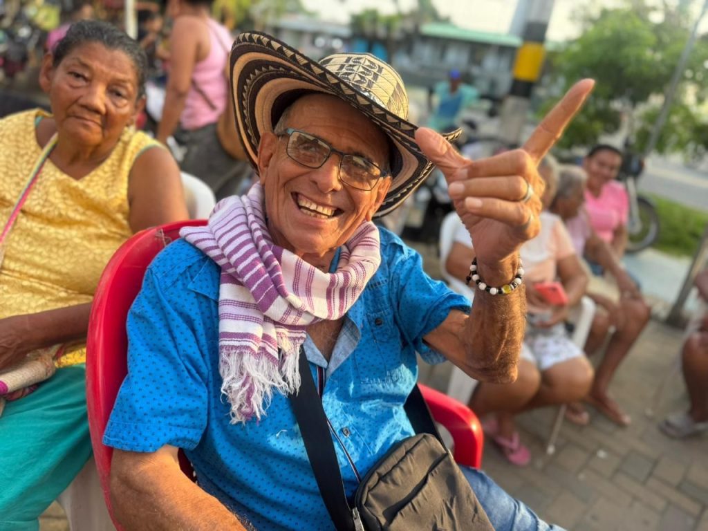 En la fotografia Un hombre mayor sonriente con sombrero vueltiao, camisa azul y pañoleta, levantando un dedo en señal de saludo, sentado junto a otras personas mayores en un espacio exterior.