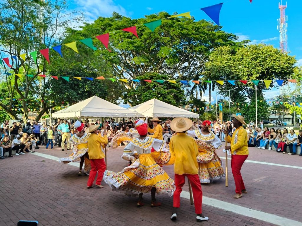 En la fotografía grupo de bailarines folclóricos con trajes típicos amarillos y rojos bailando en una plaza pública decorada con banderines de colores ante el público