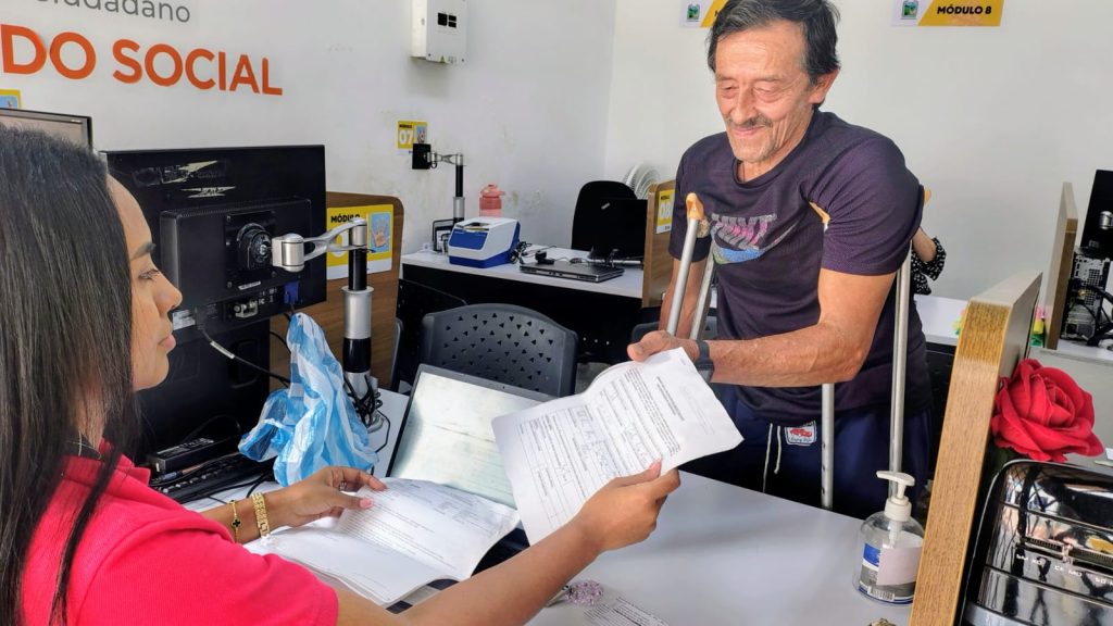 En la fotografía una persona en ventanilla atiende a un usuario que se apoya en muletas y le entrega unos documentos impresos en un módulo de atención social.