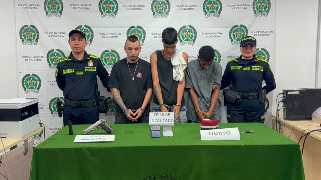 Fotografía de una mesa de evidencias de la Policía Nacional de Colombia con elementos incautados. Detrás de la mesa se encuentran dos integrantes de la policía custodiando a tres personas bajo arresto.