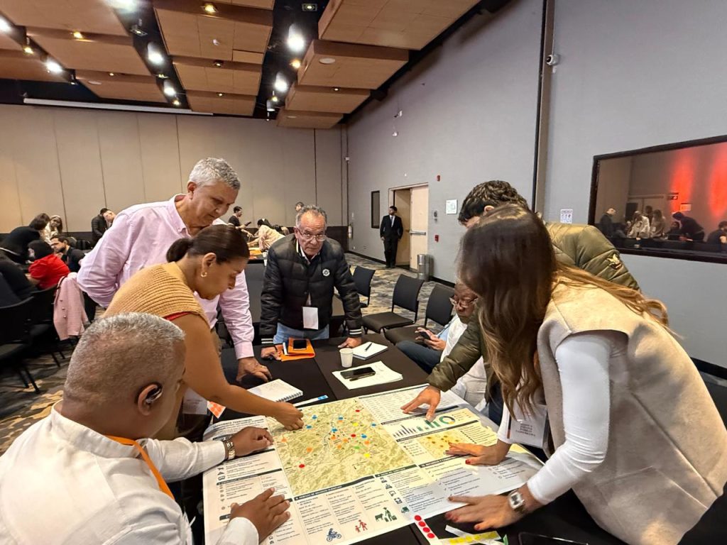 En la imagen Un grupo intergeneracional de personas se reúne alrededor de una mesa de trabajo para analizar un mapa detallado y varios documentos impresos durante un taller participativo.