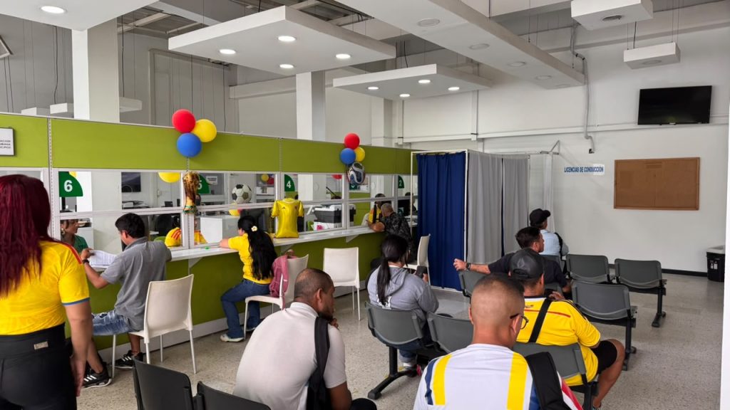 En la fotografía Vista general de una oficina de atención al ciudadano de la Secretaría de Tránsito de Palmira. Varias personas se encuentran sentadas en la sala de espera y otras son atendidas por personal administrativo en módulos con ventanillas numeradas. De fondo se aprecia decoración alusiva a la selección colombiana de fútbol.