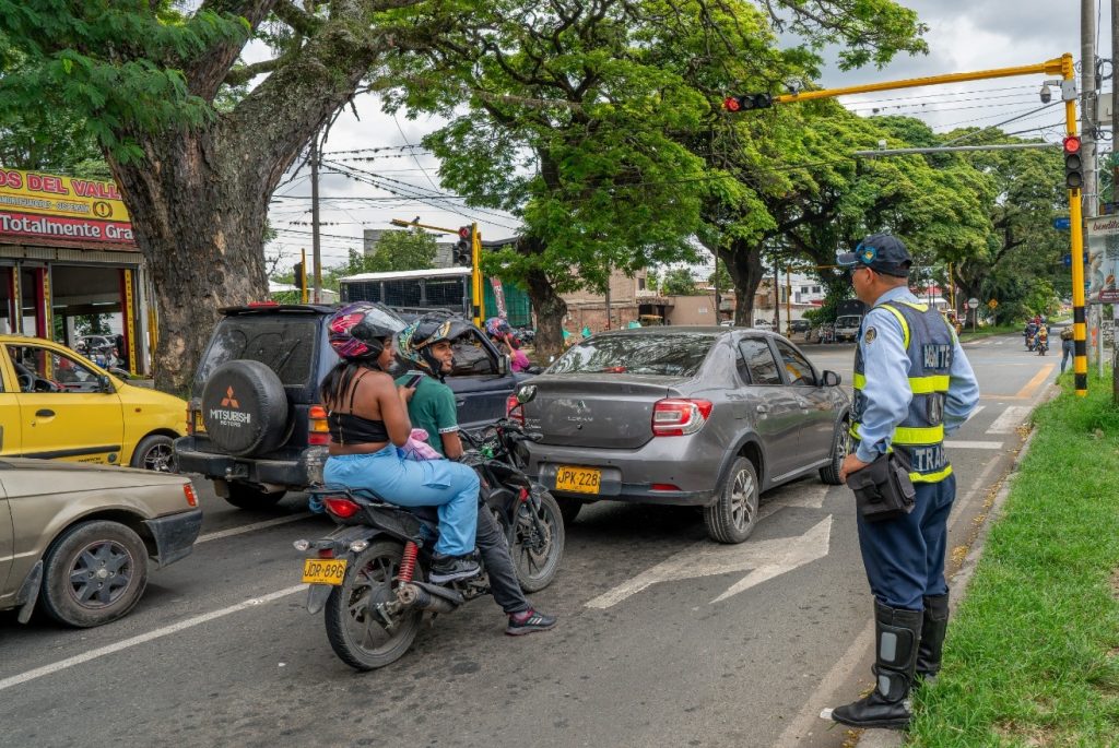 En la fotografía un agente de la Secretaría de Tránsito y Transporte de Palmira realiza labores de control y regulación del tráfico en una vía urbana congestionada. A su izquierda, se observan automóviles y una motocicleta detenidos en el flujo vehicular.