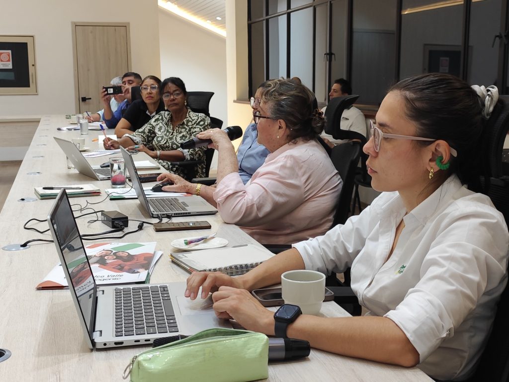 En la imagen una mesa de reuniones donde varias personas participan en una sesión de planificación turística. En primer plano, una mujer redacta en su computadora portátil, mientras que más adelante otra de las participantes interviene hablando por un micrófono de mano.
