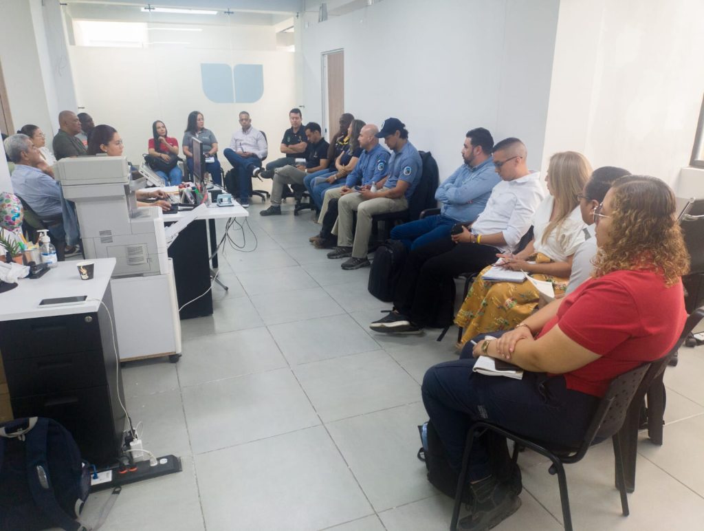 En fotografía un grupo de personas asiste a una reunión de trabajo o capacitación en una oficina luminosa, sentadas en sillas dispuestas en semicírculo mientras escuchan una presentación