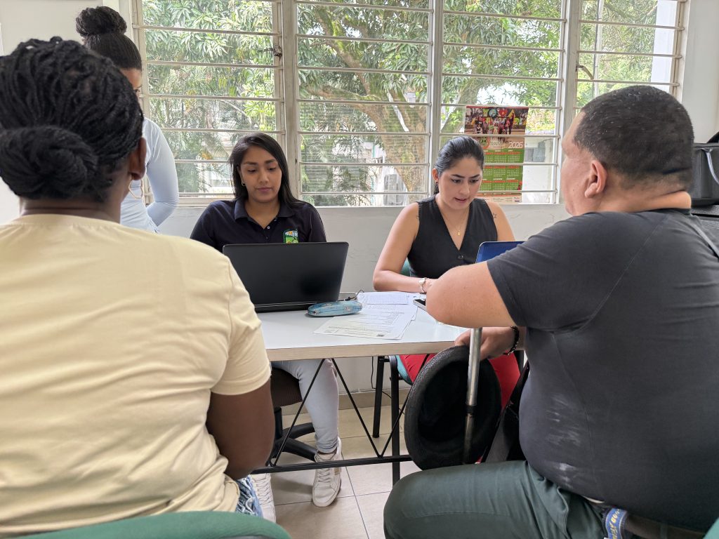 En la fotografía un grupo de personas se reúne alrededor de una mesa de oficina para una consulta o atención ciudadana