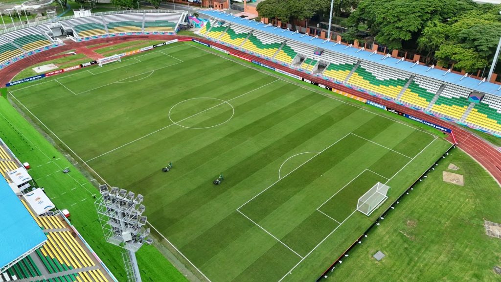 En la fotografía Vista aérea panorámica de un campo de fútbol profesional verde y demarcado, rodeado por tribunas con asientos de colores verde, amarillo y azul, y una pista de atletismo perimetral