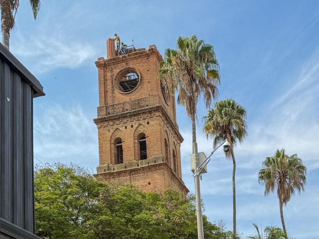Una imagen de una torre de reloj de ladrillo antiguo con personas trabajando en la parte superior, rodeada de palmeras.