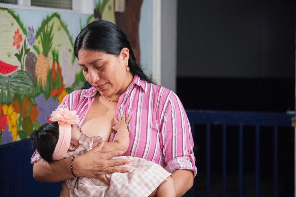 En la fotografía una persona amamanta a un bebé con un lazo rosa en la cabeza, sosteniéndole en brazos en un entorno decorado con un mural infantil.