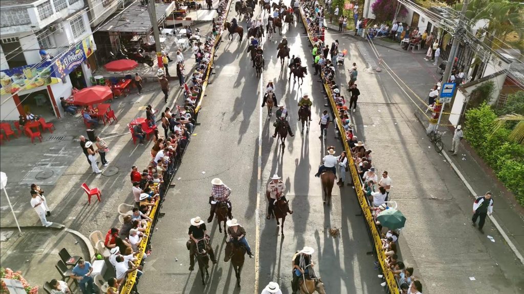 Vista aérea de un desfile de jinetes a caballo avanzando por una calle bordeada por multitudes detrás de vallas amarillas