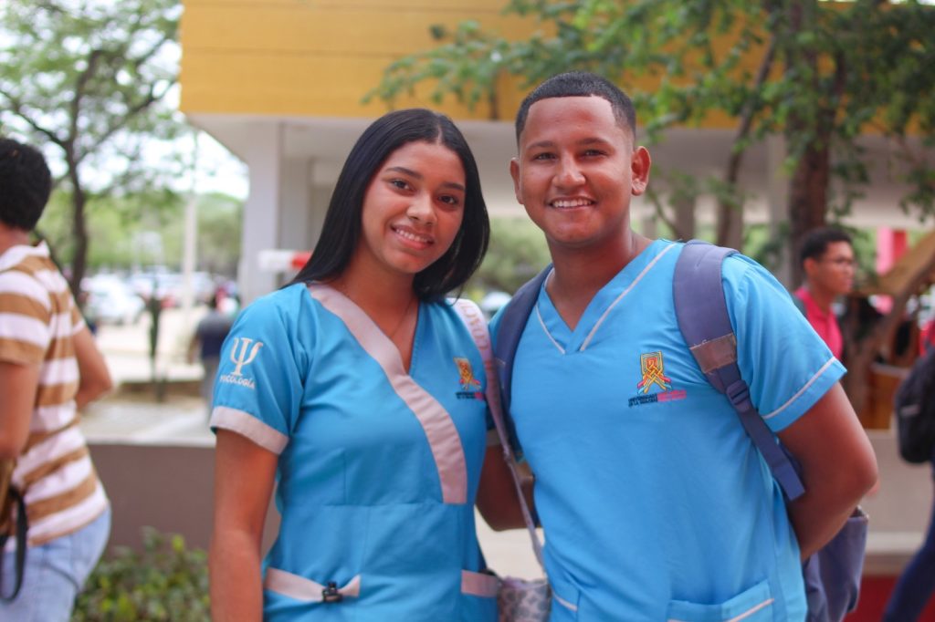 En la fotografía Dos estudiantes de Psicología de la Universidad de La Guajira sonriendo en el campus universitario con sus uniformes médicos azules.