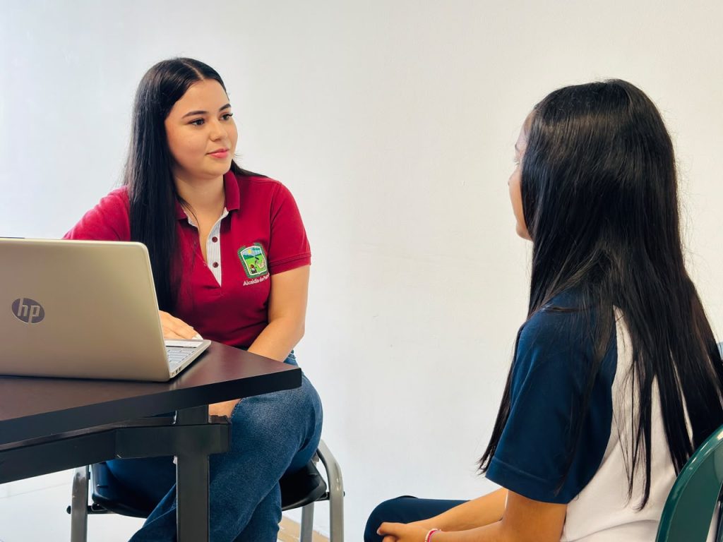 Una orientadora o profesional con camiseta institucional escucha de manera atenta a una estudiante durante una sesión individual de acompañamiento.