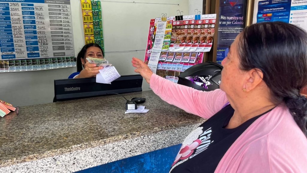 En la fotografía una empleada de un punto de atención de servicios de giros y lotería entrega dinero y un recibo a una usuaria en la ventanilla.