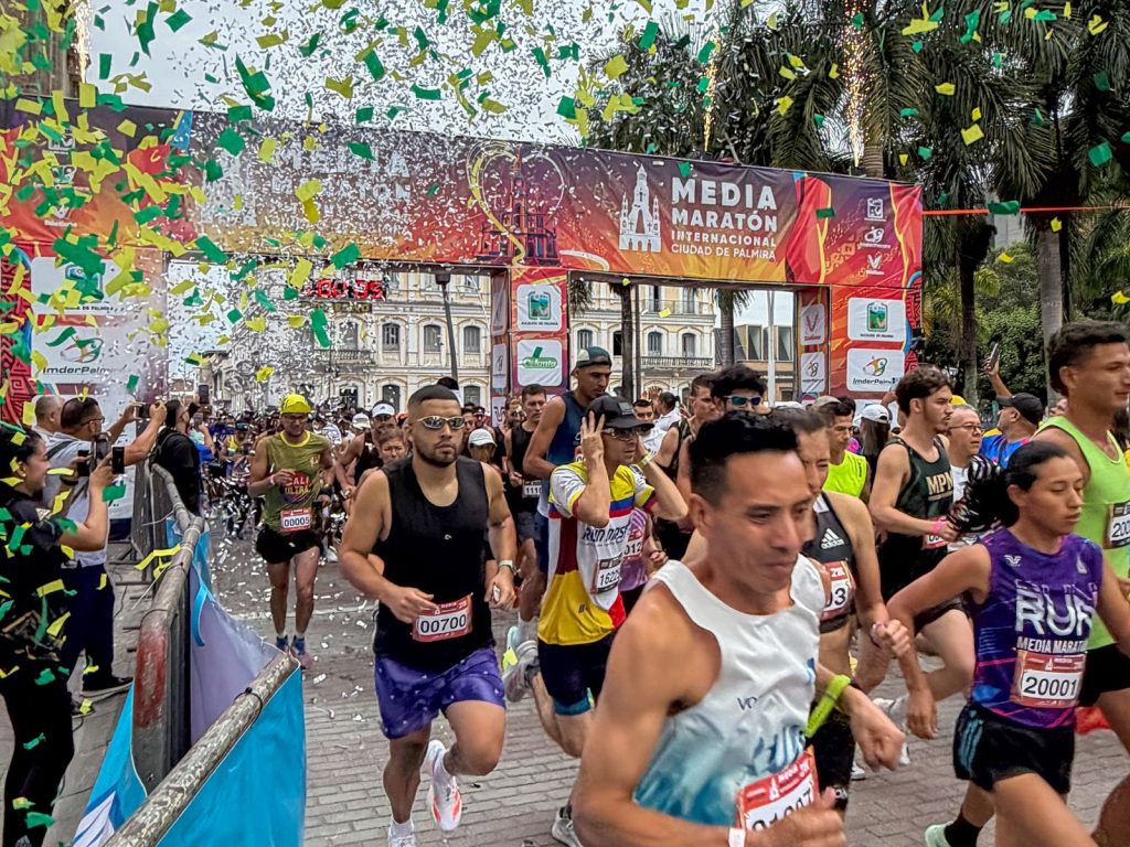 Grupo de corredores en el punto de salida de la Media Maratón Internacional Ciudad de Palmira, bajo una lluvia de confeti verde y amarillo frente al arco oficial del evento.