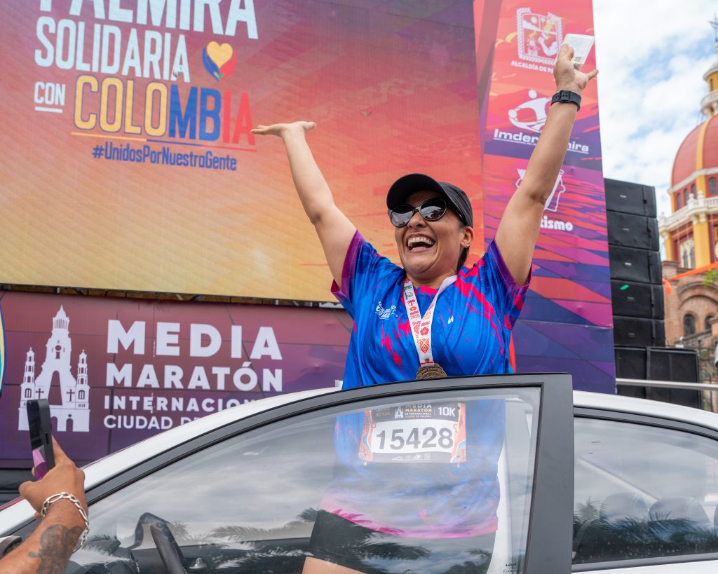 Mujer ganadora sonriente con medalla al cuello y gorra negra levantando los brazos en señal de victoria desde la puerta de un vehículo blanco, tras participar en la Media Maratón Internacional Ciudad de Palmira.