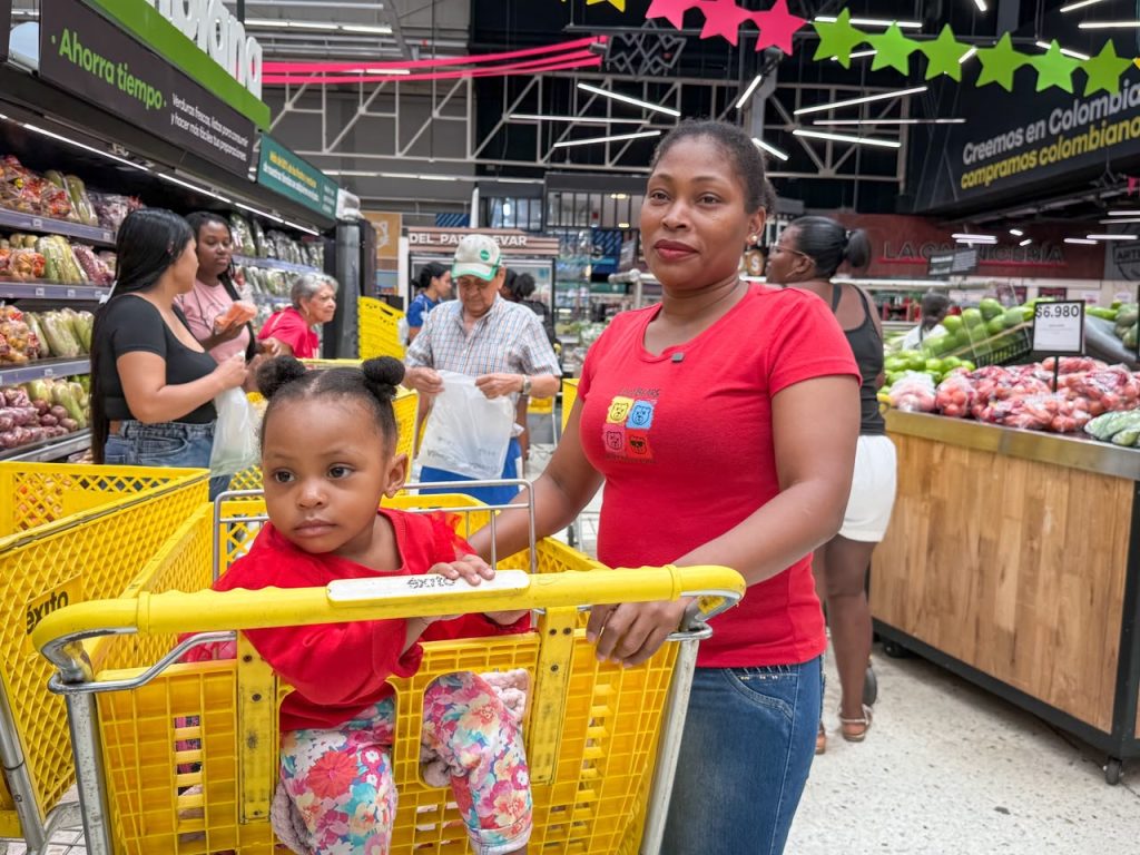 En la fotografía Una mujer y una niña pequeña con camisetas rojas sonríen mientras compran juntas en la sección de frutas y verduras de un supermercado.