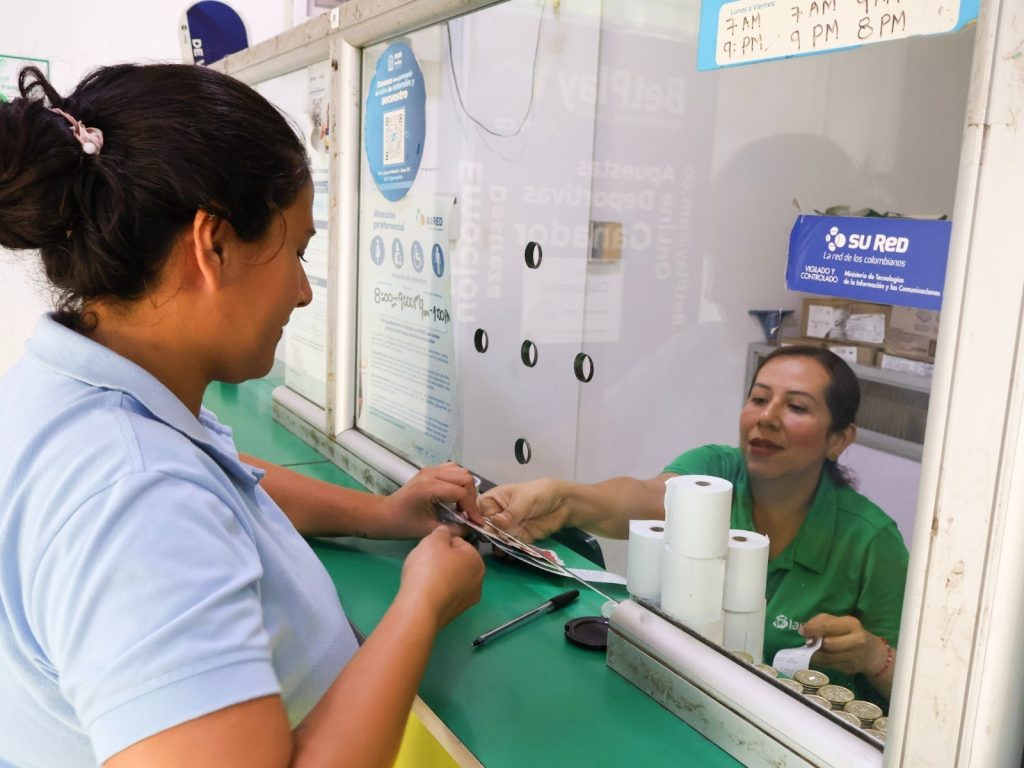 En la imagen Una cajera atiende a una clienta a través de una ventanilla de servicio en un punto de atención de SuRed.