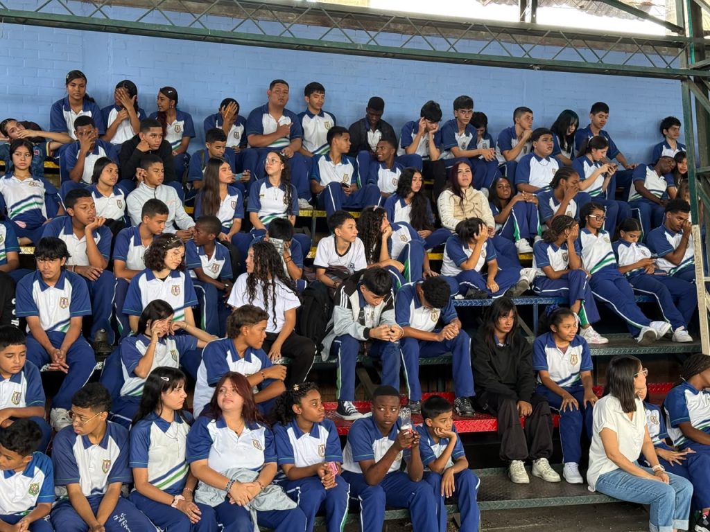 Grupo de estudiantes con uniformes escolares azules y blancos sentados en las graderías de un polideportivo durante un evento de la Semana Andina.