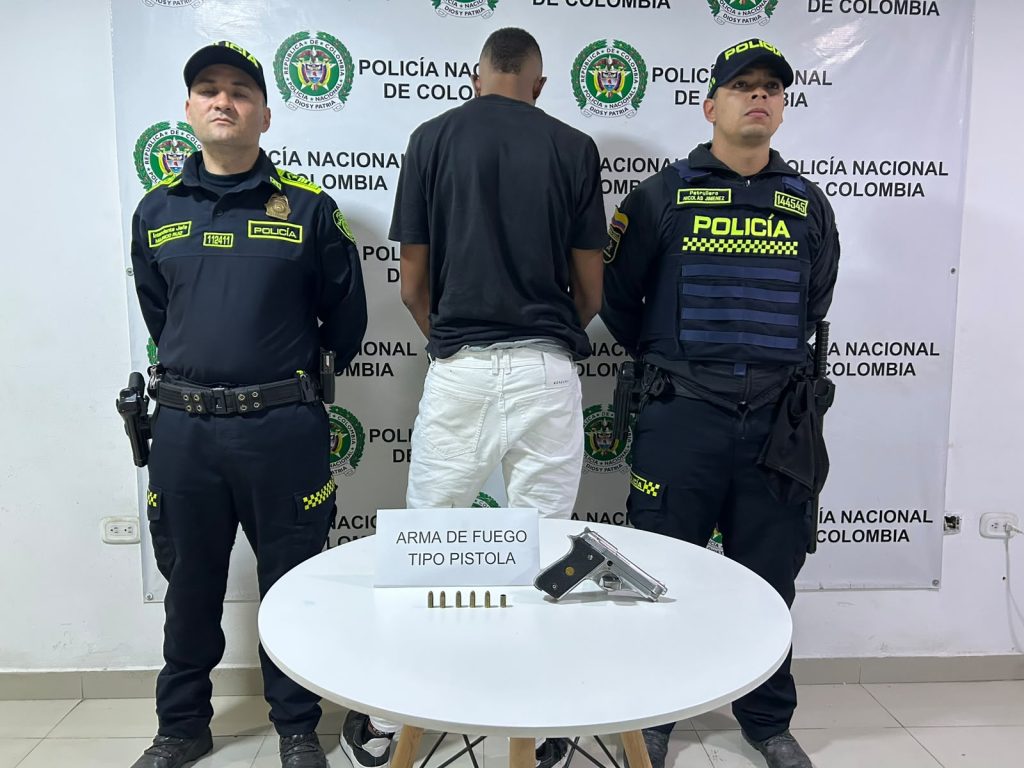 En la foto Dos integrantes de la Policía Nacional de Colombia custodian a una persona detenida que se encuentra de espaldas. Delante, sobre una mesa, se exhiben una pistola y seis cartuchos incautados