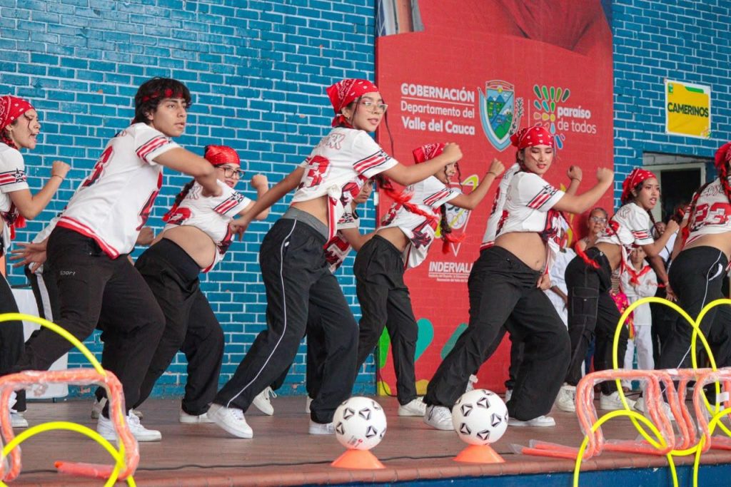 En la imagen un Grupo de jóvenes con pañoletas rojas y ropa deportiva blanca y negra realizan una coreografía de baile urbano en una tarima.