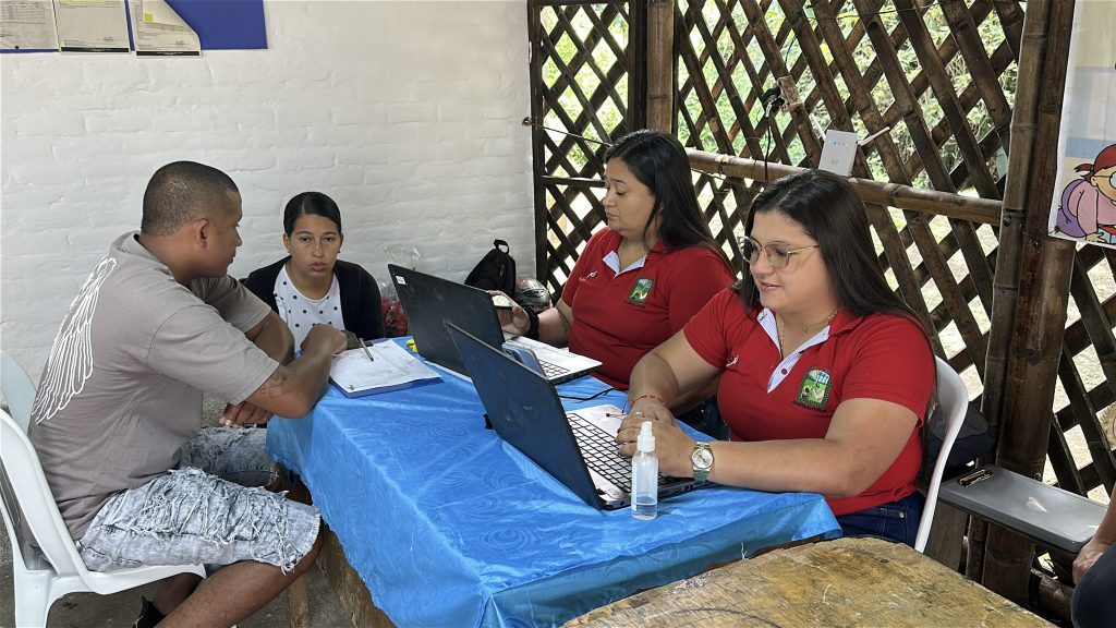 En la imagen dos personas de la organización, que visten camisas rojas, atienden a una pareja con computadoras portátiles en una mesa de registro.