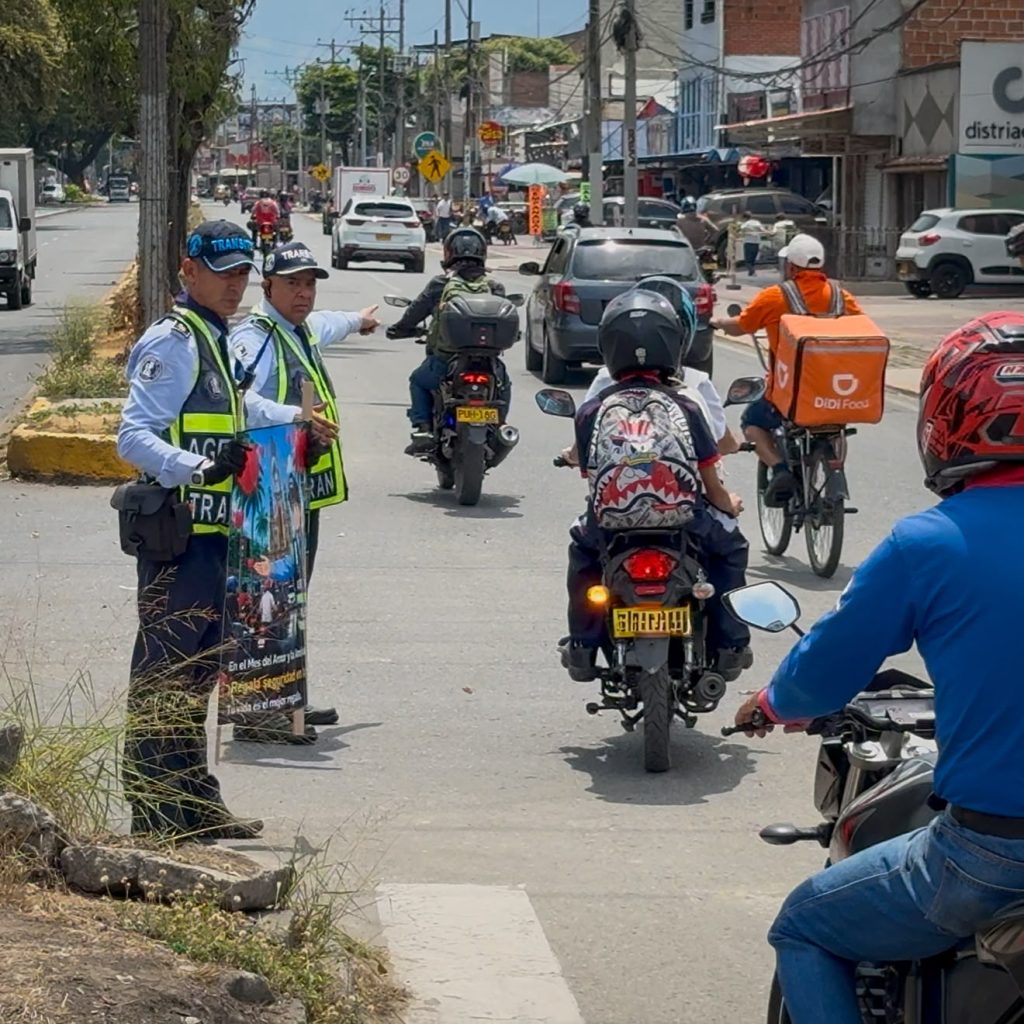 Fotografía en una vía urbana donde dos agentes de tránsito sostienen un cartel pedagógico y orientan el flujo vehicular, interactuando con personas que se desplazan en motocicletas y bicicletas.
