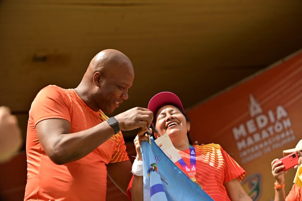 En la foto alcalde de Palmira con camiseta naranja le coloca una medalla a otra persona que sonríe con alegría portando una gorra rosa y sosteniendo una bandera celeste, durante la premiación de la Media Maratón Internacional