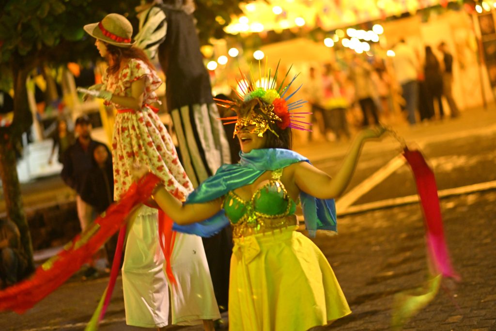 En la foto una artista con un colorido tocado de picos, antifaz dorado y falda amarilla baila con cintas rojas en un desfile callejero nocturno junto a dos personas en zancos