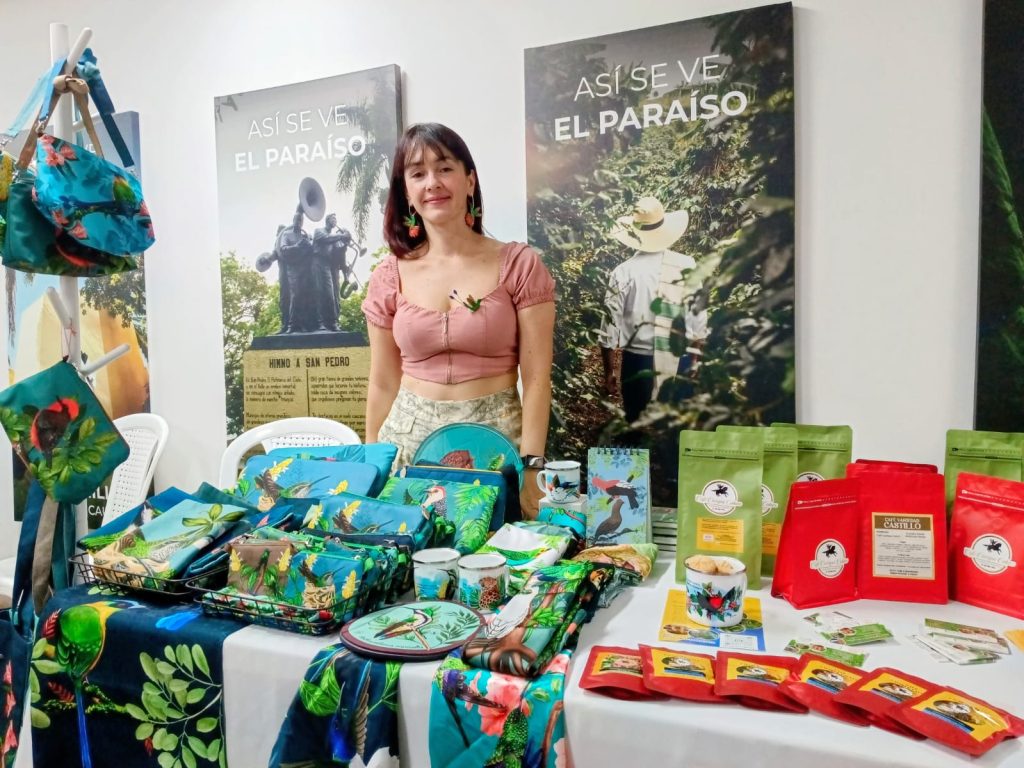 Una persona emprendedora posa sonriente detrás de un stand de productos artesanales y café local, bajo carteles con la frase "Así se ve el paraíso".