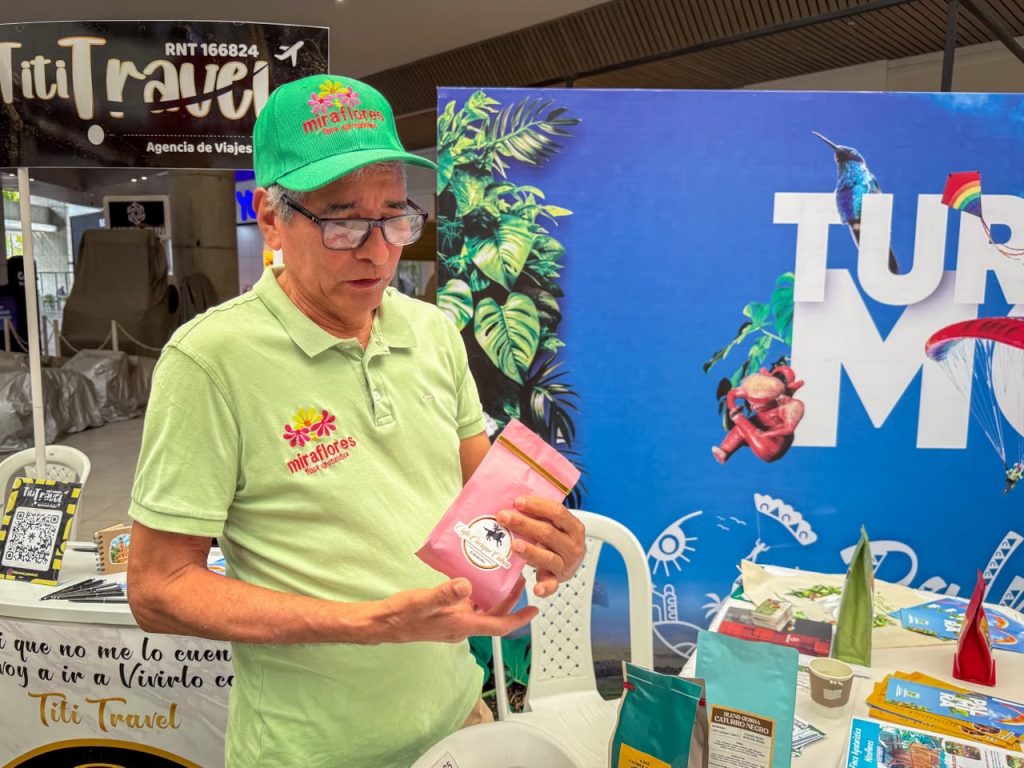 En la foto hombre con gorra verde y polo de Miraflores mostrando un paquete de café rosa en una feria de turismo