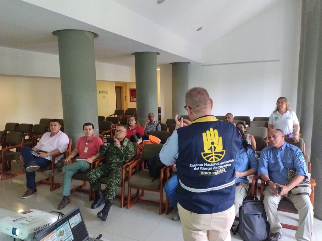 En la foto Una persona de espaldas con chaleco del Sistema Nacional de Gestión del Riesgo de Desastres (DGRD Palmira) se dirige a un grupo de personas sentadas en un auditorio durante una capacitación.