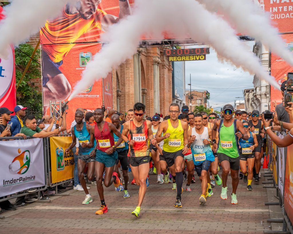 En la fotografía Grupo de atletas de alto rendimiento e ingenio deportivo arrancan la carrera atravesando la arquería de salida entre columnas de humo blanco durante la Media Maratón de Palmira.