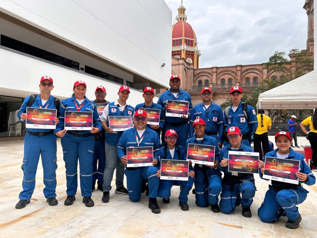 Grupo de voluntarios de la Cruz Roja con uniformes azules y gorras rojas posando en una plaza exterior mientras sostienen diplomas de reconocimiento de la Media Maratón Internacional Ciudad de Palmira.