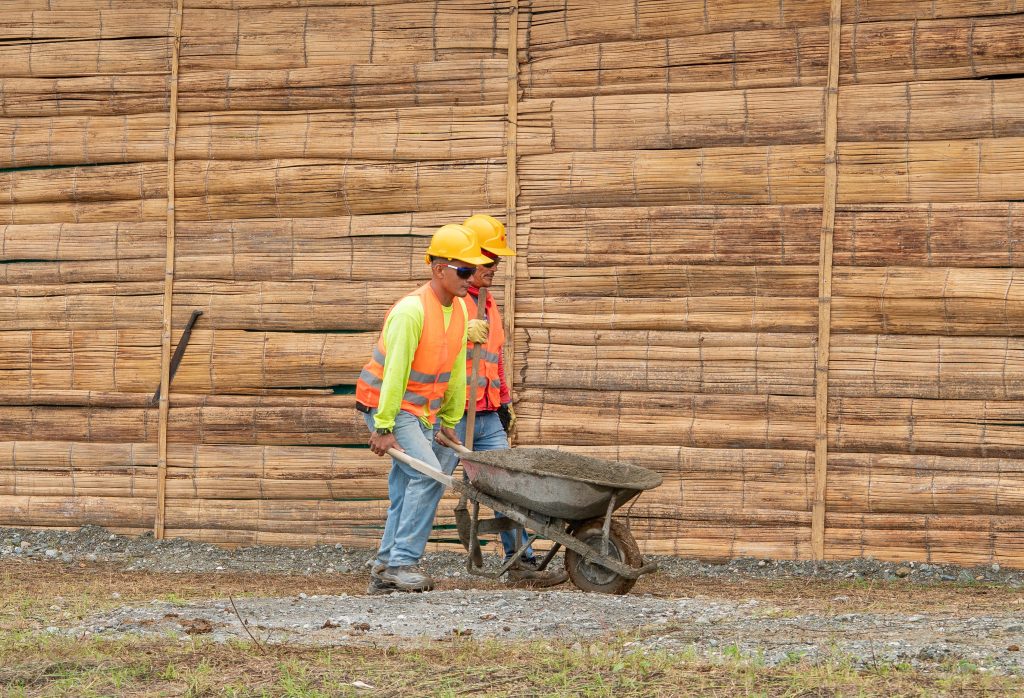 Dos personas del equipo de construcción transportan material en una carretilla vistiendo cascos y chalecos de seguridad frente a una pared de caña o guadua.